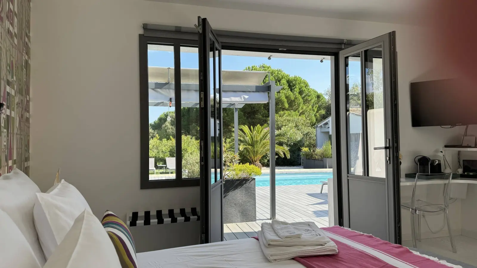 Bedroom at Le 36 with French windows open onto the wooden deck and pool, towels laid on the bed