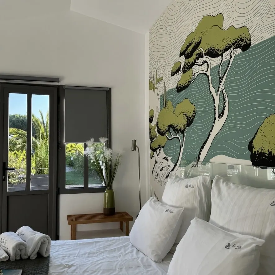 Bedroom at Le 36 with pine-and-sea wallpaper, white bed, towels and a glazed door to the garden
