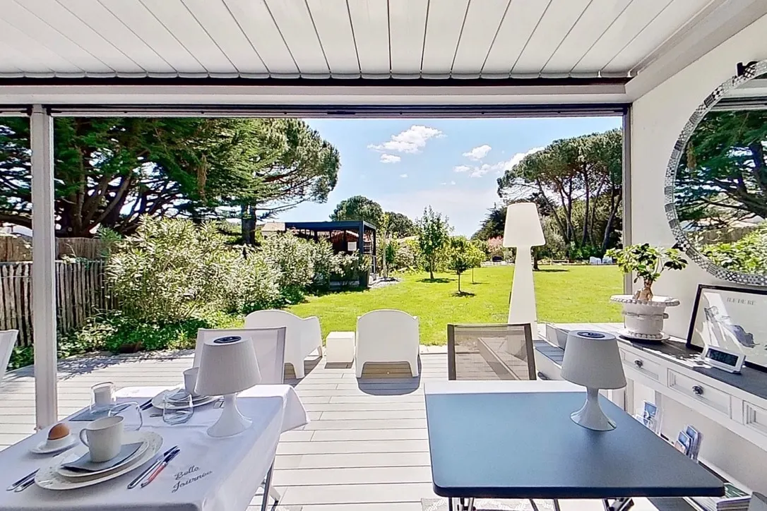 Breakfast area at Le 36 under a white pergola, laid tables opening onto the lawn and pine trees