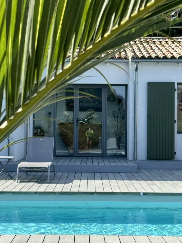 White facade of Le 36 with glass doors, green shutter and a sun lounger on the wooden deck by the pool