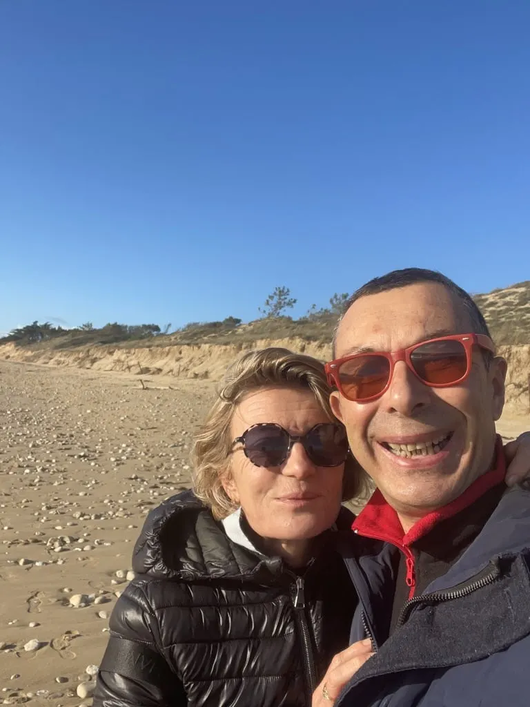 The hosts of Le 36, a smiling couple in sunglasses, taking a selfie on a sandy beach below the dunes