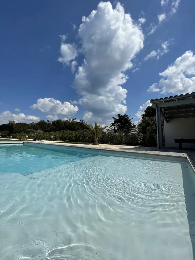 Clear water of the pool at Le 36 under a blue sky with big white clouds, pool house and palm behind
