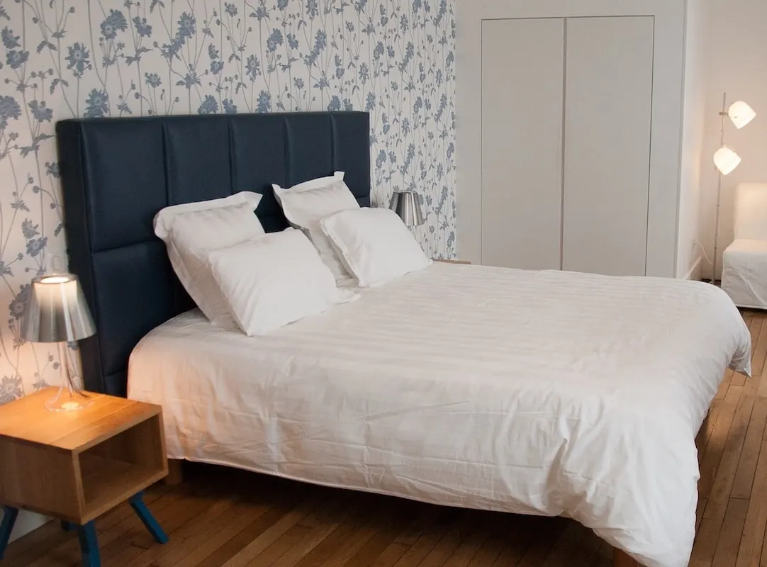 Chambre de La Maison M : lit blanc, tête de lit capitonnée bleu nuit, papier peint floral et parquet