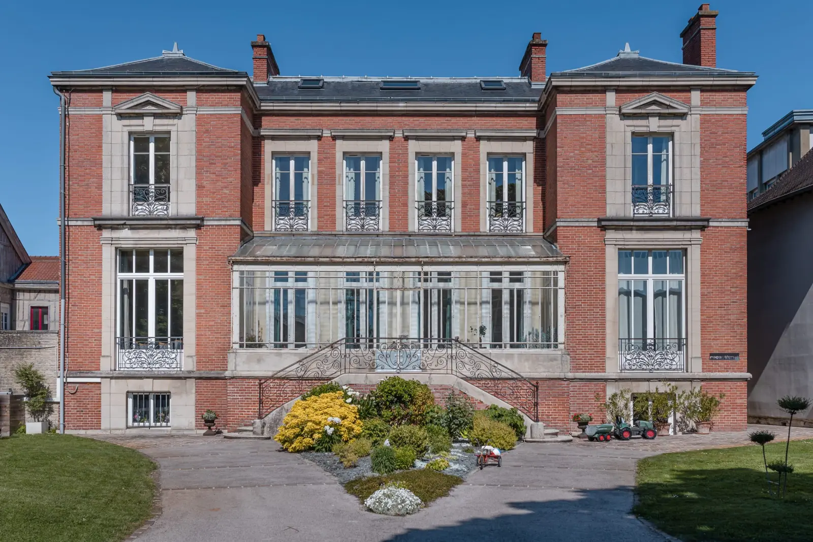 Façade d'une maison de maître en brique rouge avec véranda, escalier en fer forgé et massifs fleuris