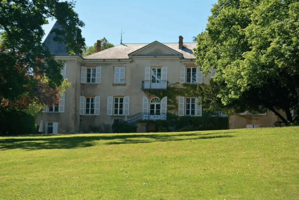 Manor house with white shutters and a wrought-iron balcony at the end of a wide lawn ringed with trees