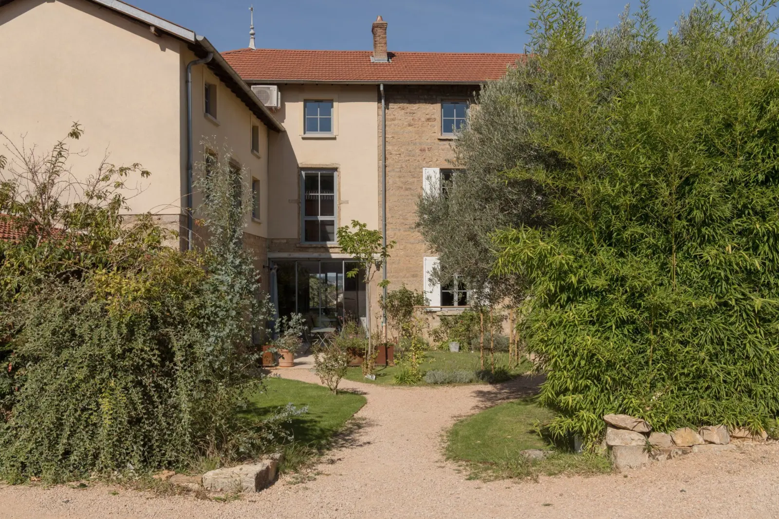 Stone and pale rendered buildings at the end of a gravel drive lined with greenery, La Maison des Monts d'Or