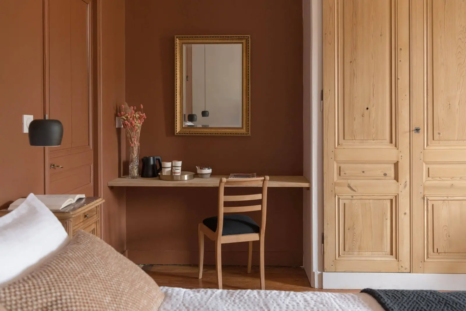 Desk, wooden chair and gilt mirror against a terracotta wall, pale wooden cupboard, La Maison des Monts d'Or
