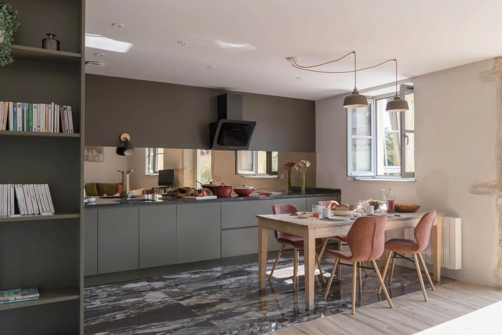 Open-plan gîte kitchen with a table, pink chairs, marbled floor and bookshelves, La Maison des Monts d'Or