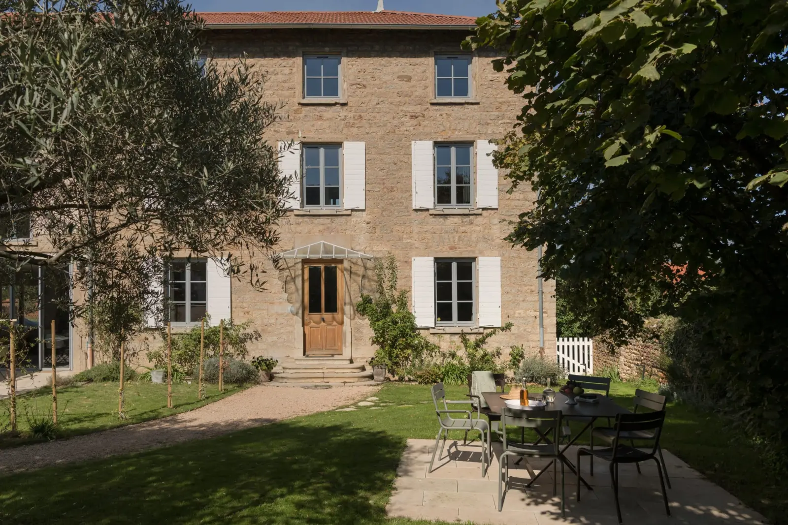 Stone façade with white shutters and a wooden door, table and chairs in the garden, La Maison des Monts d'Or