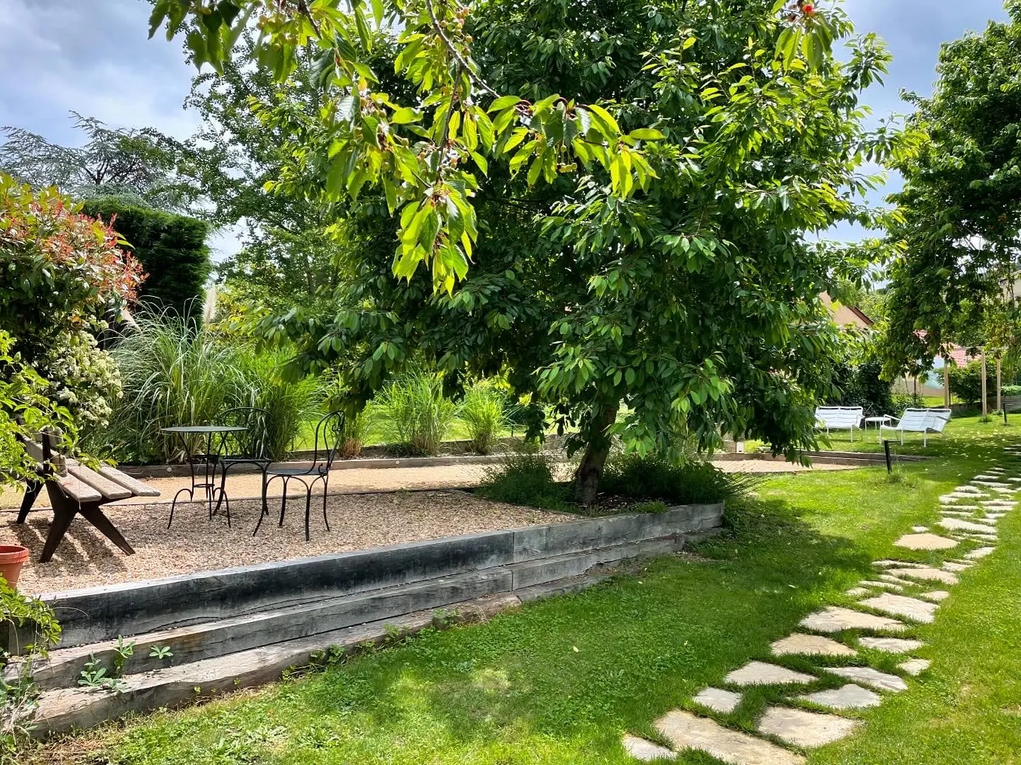 Tree-filled garden at La Maison des Monts d'Or with stepping stones, a bench and a bistro table on gravel