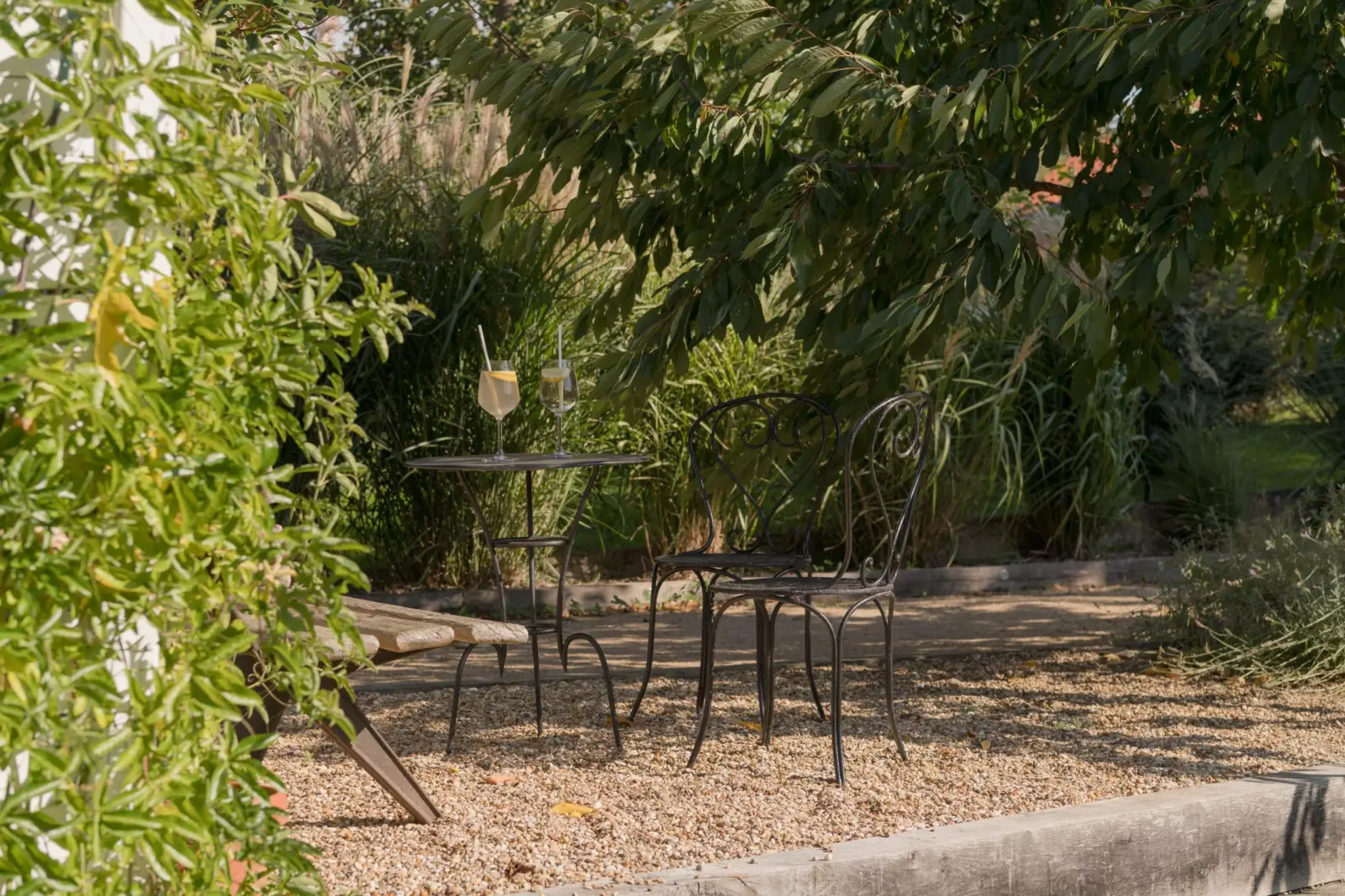 Wrought-iron bistro table with two glasses and chairs on gravel beneath the foliage, La Maison des Monts d'Or