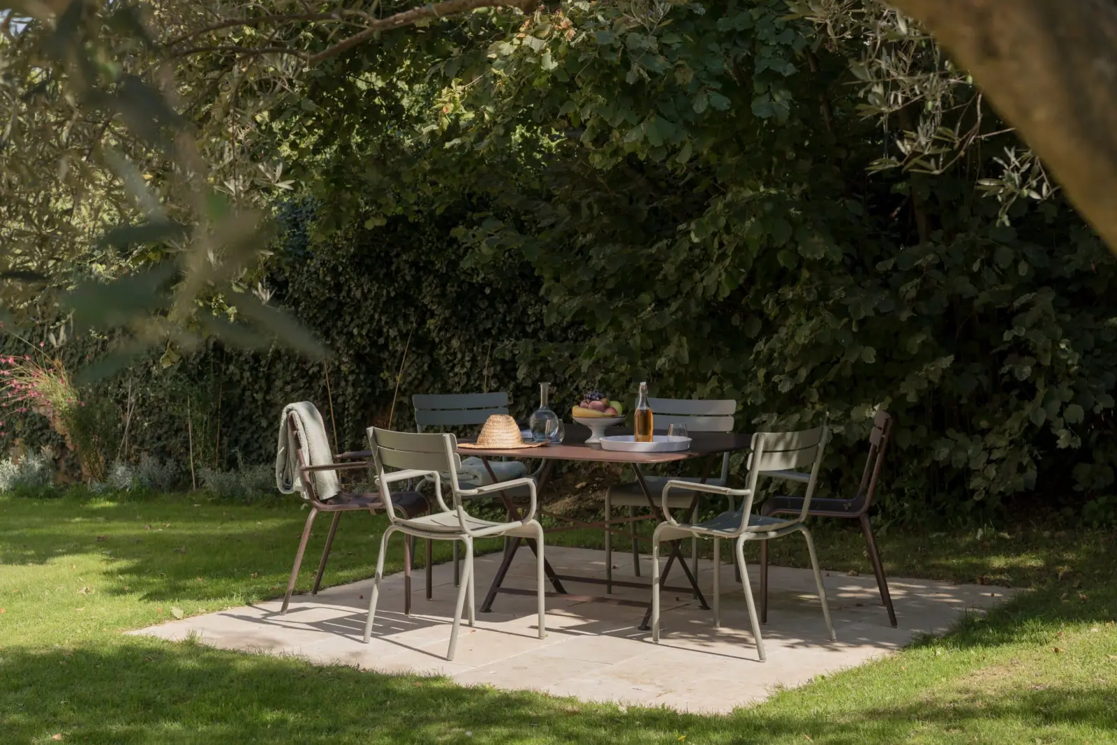 Grey-green garden table and chairs on a paved slab in the shade of trees, La Maison des Monts d'Or