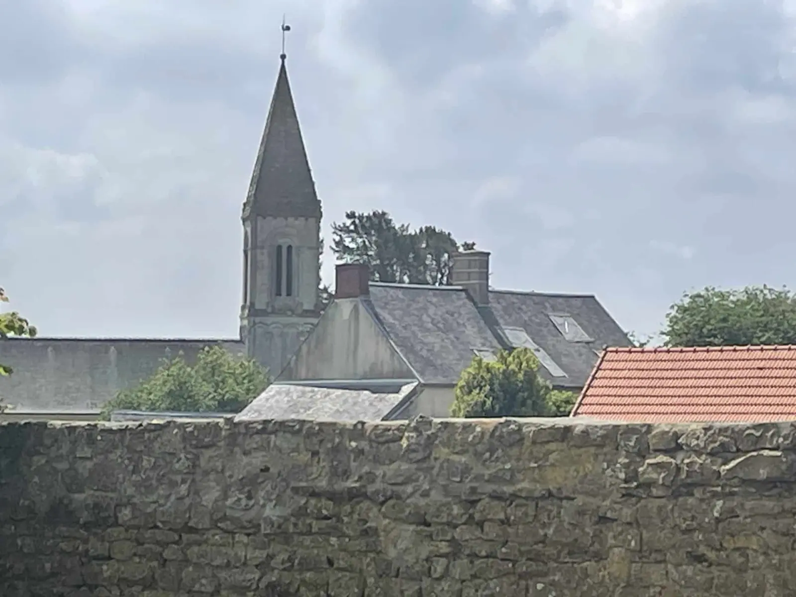 Pointed church spire above a stone wall and slate and tiled roofs, seen from Domaine de Marculfo