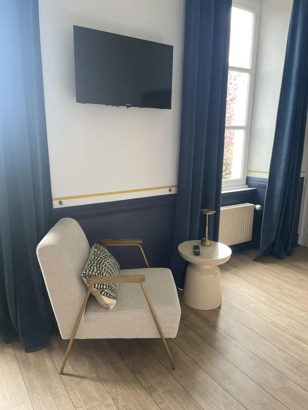 Sitting corner of the Houlgate room at Marculfo, beige armchair, side table, television and blue curtains