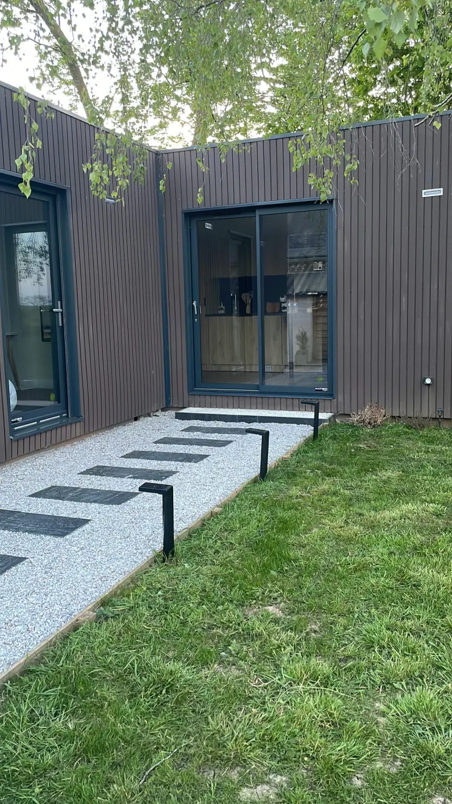 Dark timber-clad lodge at Domaine de Marculfo, glass door, gravel stepping-stone path and lawn
