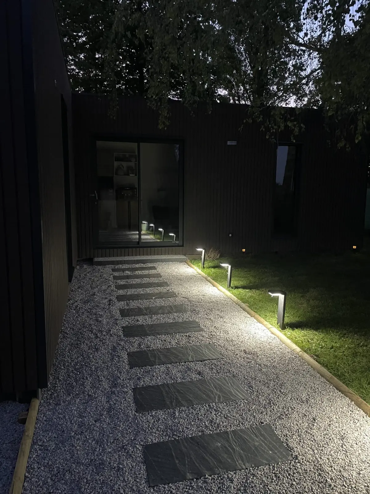 Lodge at Domaine de Marculfo at night, dark cladding, glass door and lit stepping-stone path