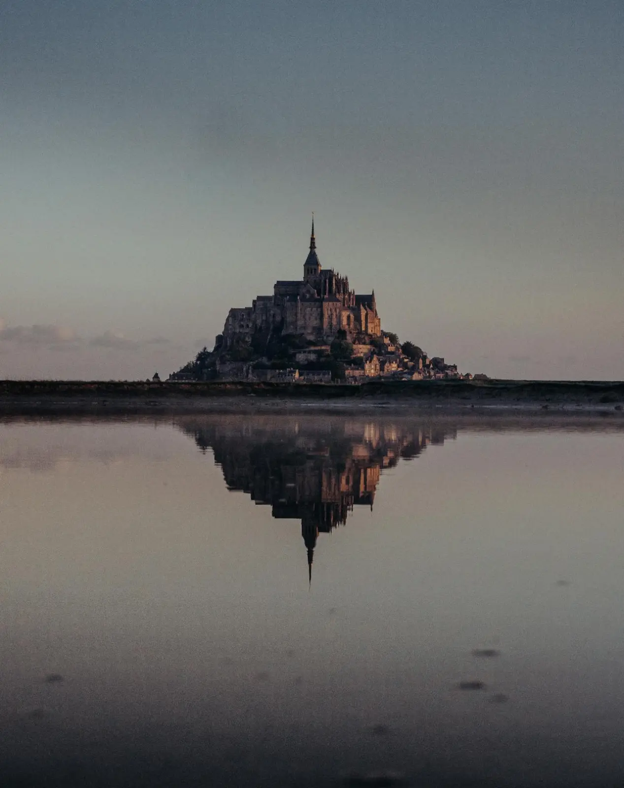 Le Mont-Saint-Michel au crépuscule, sa silhouette reflétée dans l'eau calme de la baie