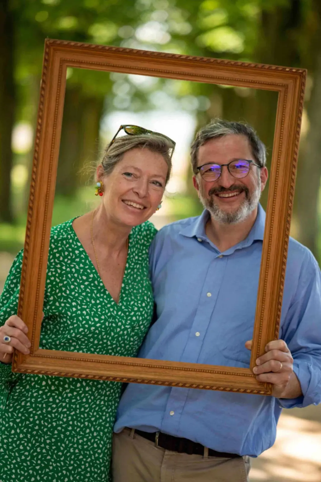 The hosts of Domaine de Montclair, smiling and holding a gilt frame in a tree-lined avenue