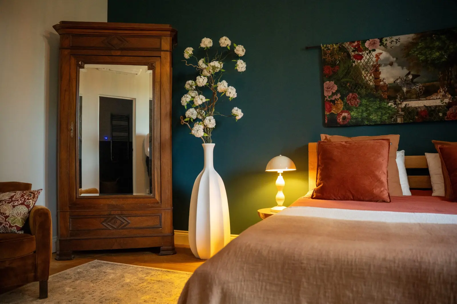 Bedroom at Le Patio Bleu with an antique mirrored wardrobe, white vase of flowers, lit lamp and dark green wall