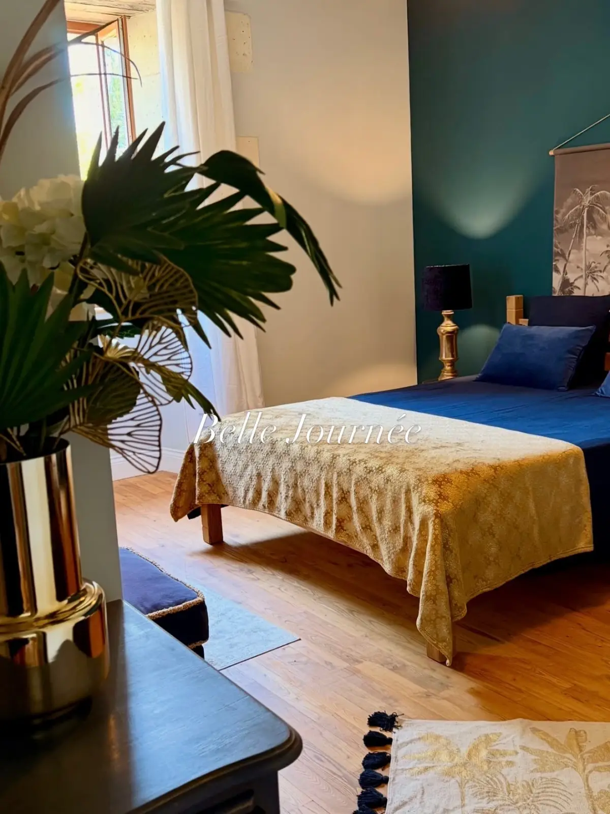 Bedroom at Le Patio Bleu with a gold bedspread on a blue bed, teal wall, palm-print hanging and plant