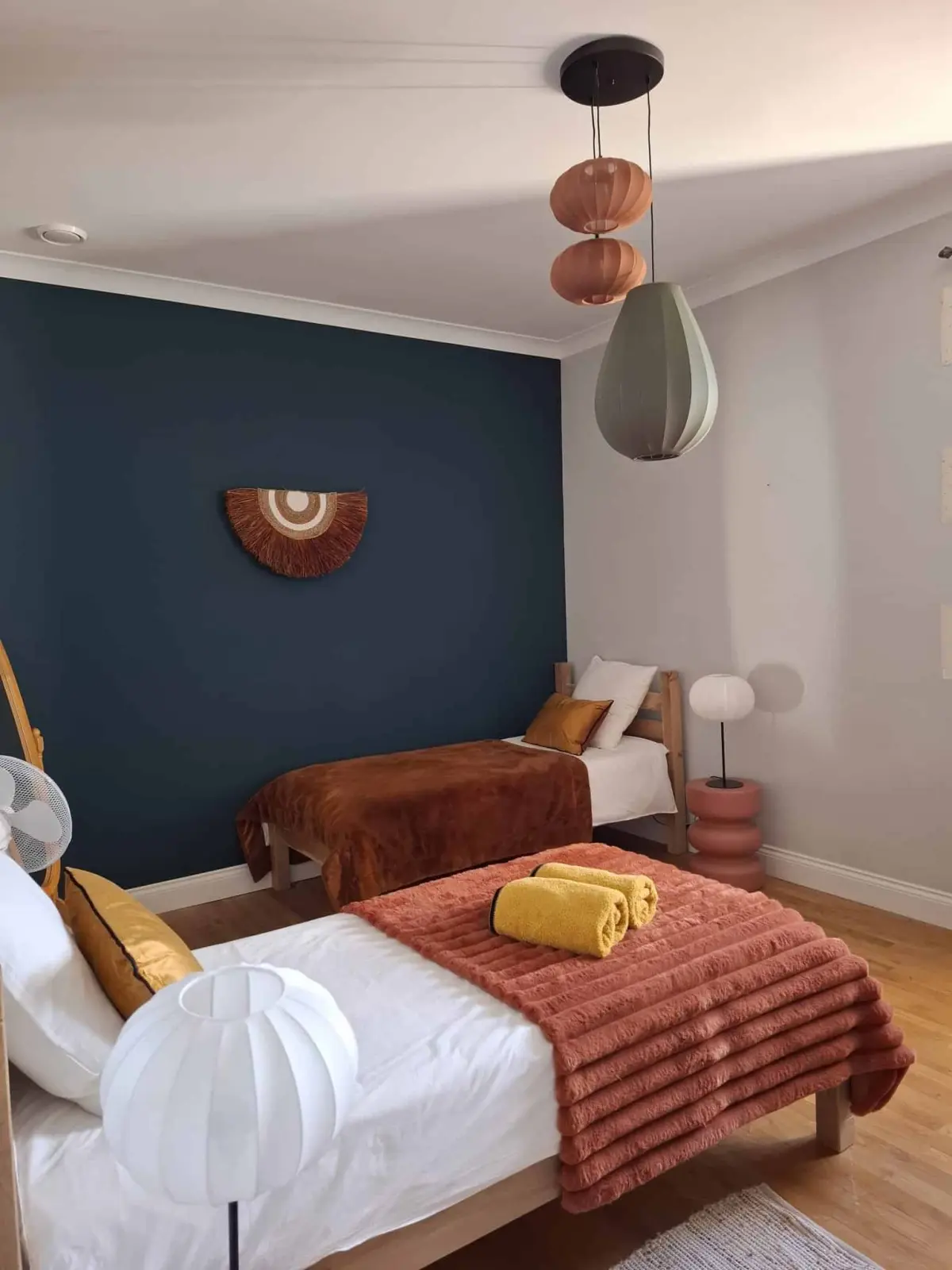 Bedroom at Le Patio Bleu with two beds, terracotta throws, yellow towels, a dark blue wall and pendant lamps