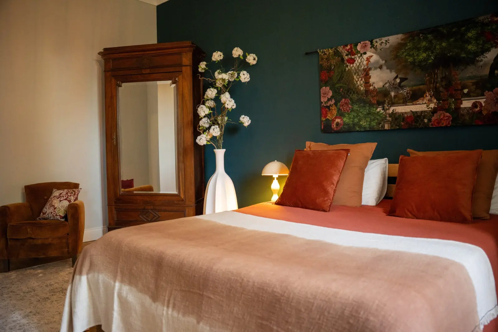 Bedroom at Le Patio Bleu with rust-coloured bedding, antique mirrored wardrobe, tall white vase and hanging