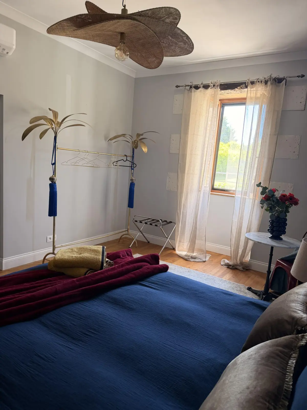 Bedroom at Le Patio Bleu with a blue bed, burgundy throw, woven fibre ceiling light, gold rails and sheer curtains