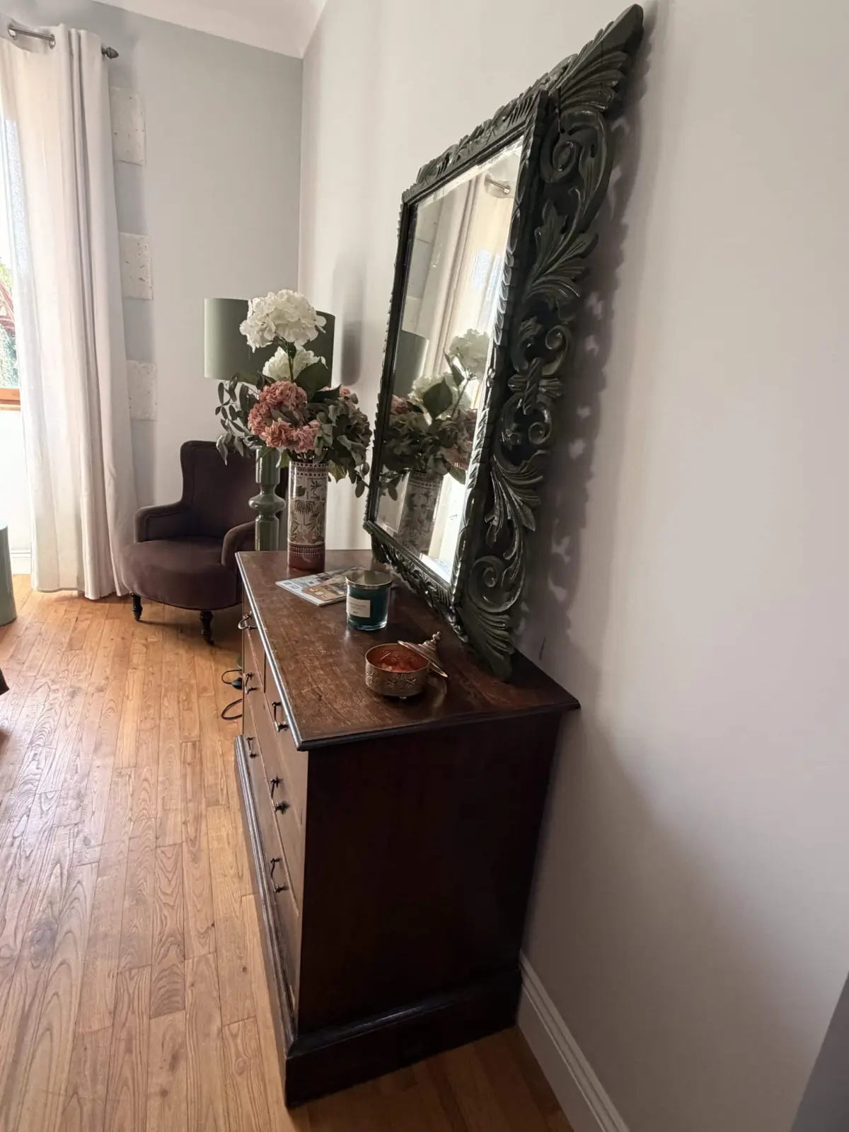 Hallway at Le Patio Bleu with a dark wood chest of drawers, large carved mirror, bouquet and armchair by the window