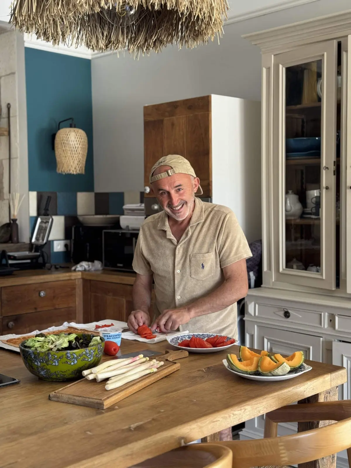 Host at Le Patio Bleu slicing tomatoes in the kitchen, with melon, salad and asparagus on the table