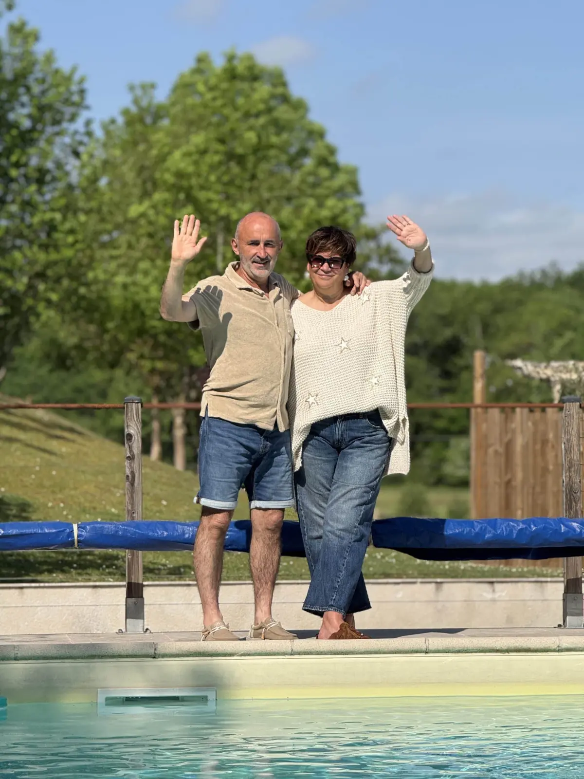 The two hosts of Le Patio Bleu waving by the pool, with trees and a blue sky behind them