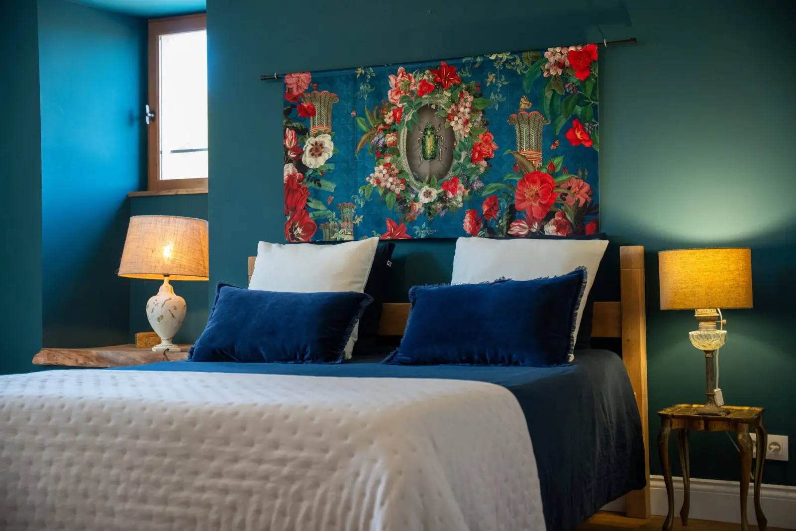 Double bed at Le Patio Bleu with blue cushions, lit bedside lamps and a floral hanging on a blue wall