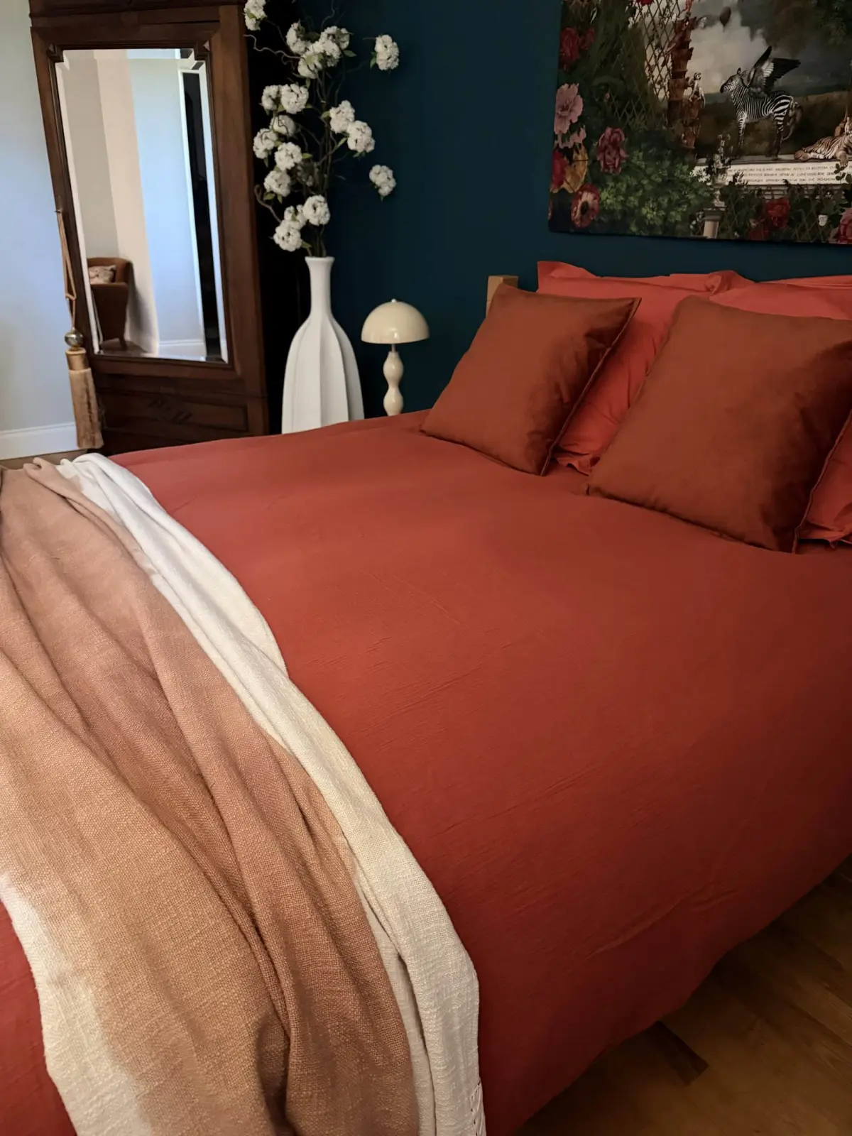 Bed at Le Patio Bleu with rust-coloured linen, pink throw, mirrored wardrobe, vase of white flowers and lamp