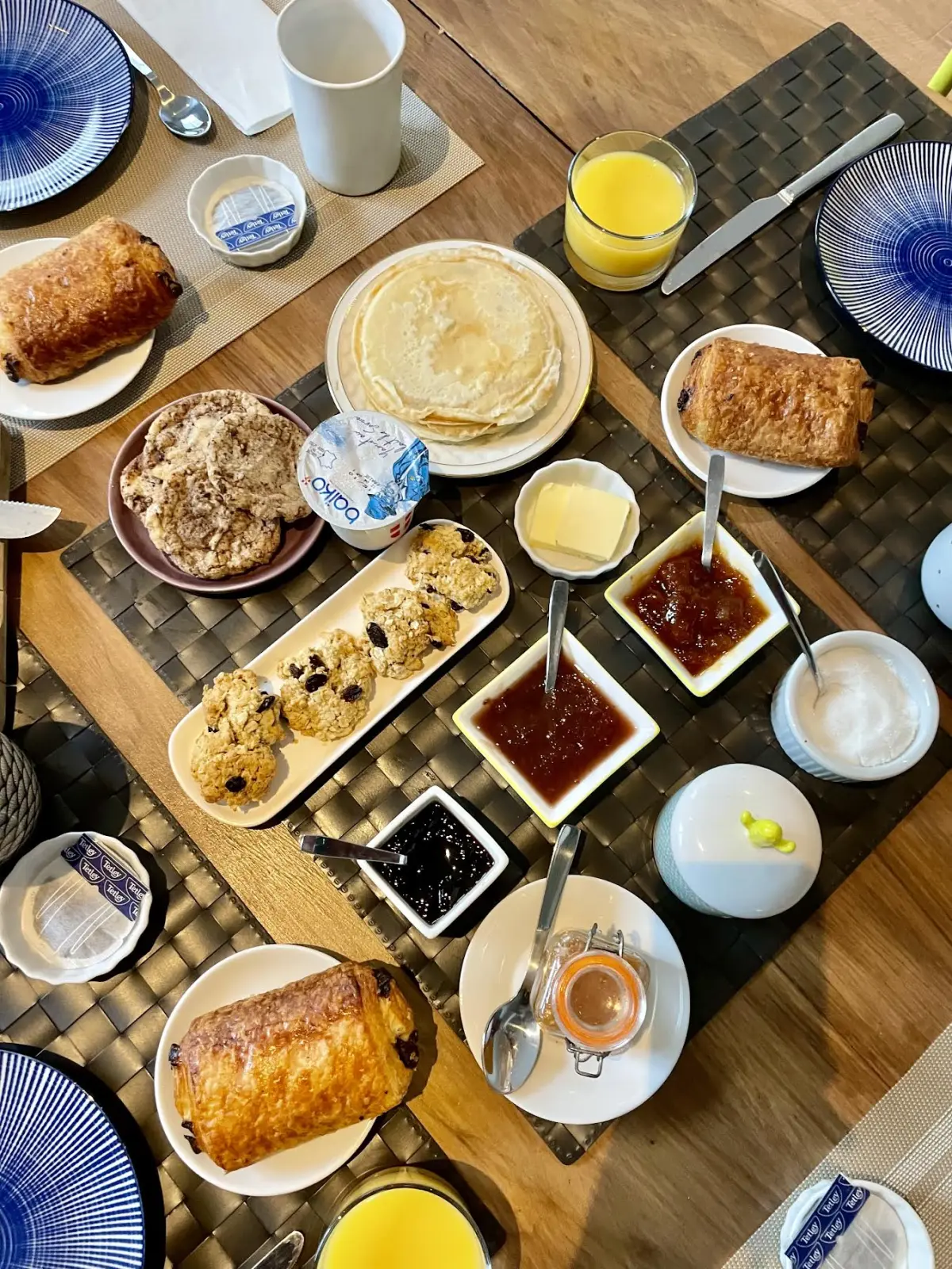 Petit-déjeuner vu du dessus : crêpes, pains au chocolat, confitures, jus d'orange et vaisselle bleue