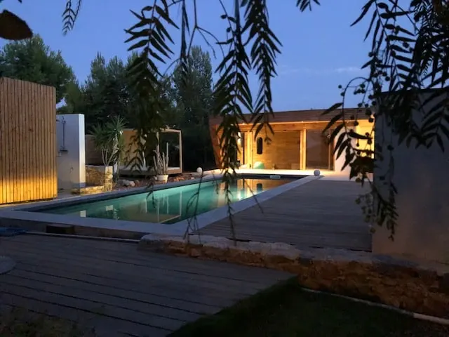 Piscine éclairée au crépuscule entre terrasse en bois et pool house, sous des feuillages