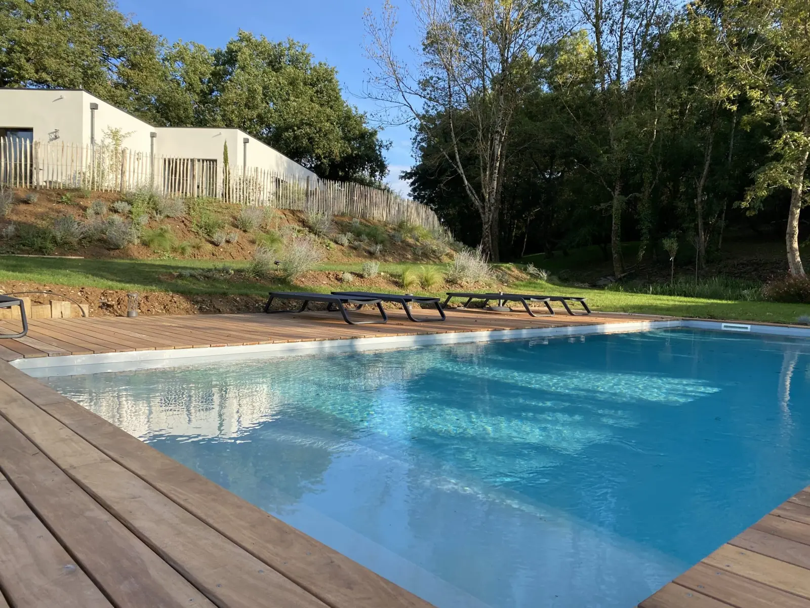Piscine à margelles en bois devant une maison blanche, transats sur la pelouse et grands arbres