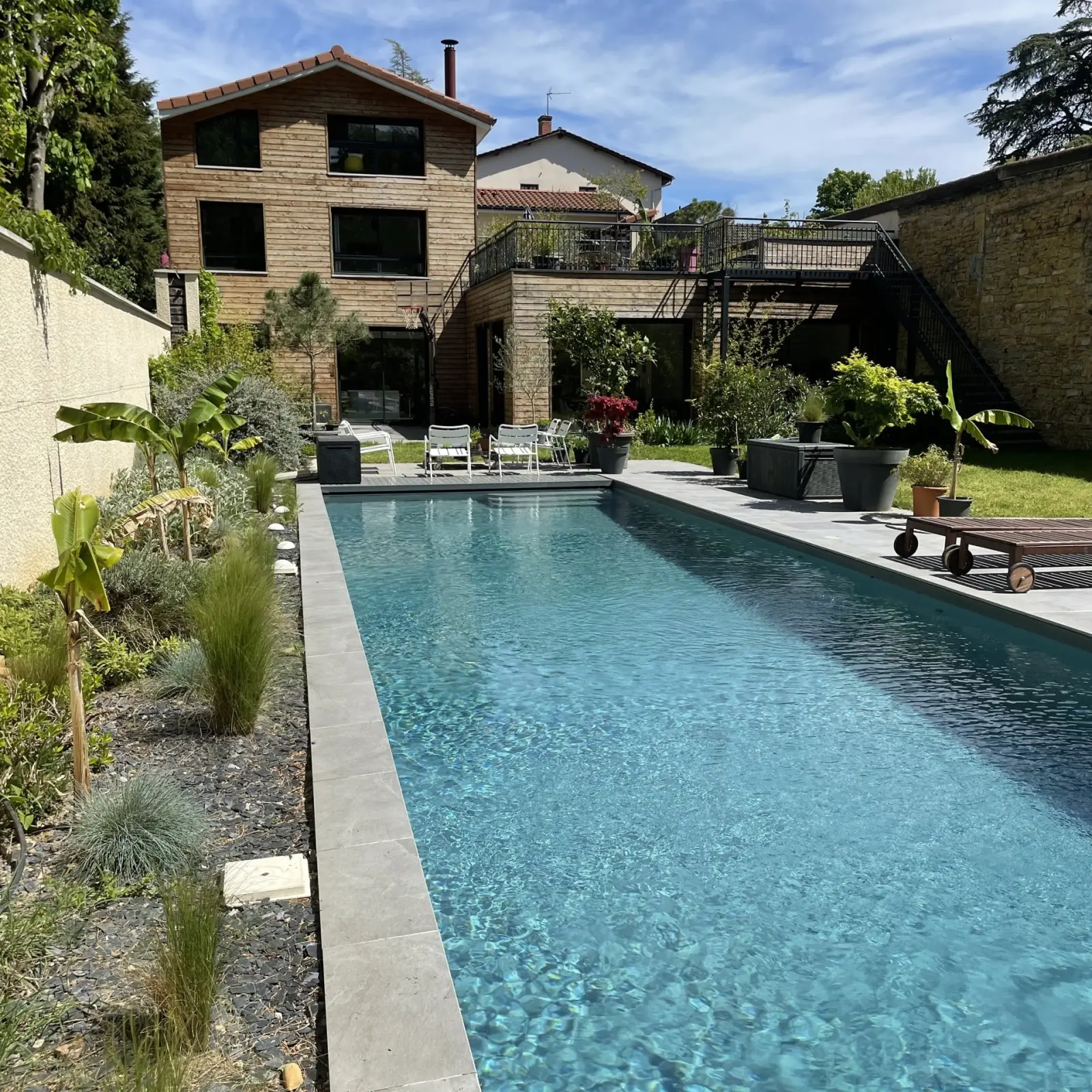 Piscine en longueur bordée de graminées devant une maison à bardage bois et sa terrasse