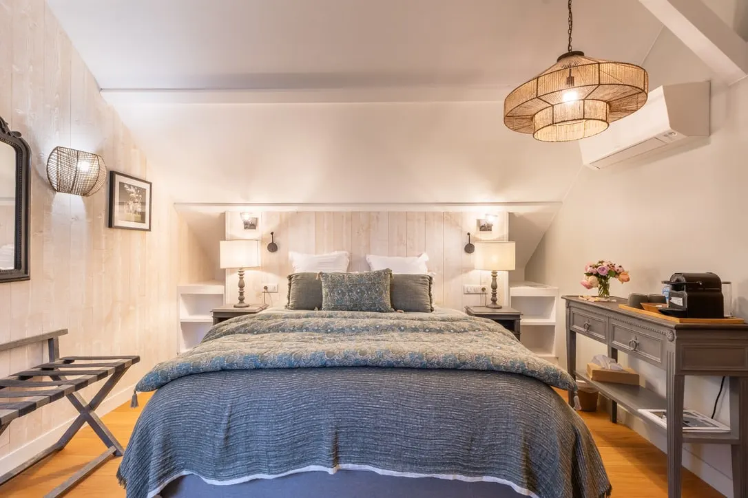 Attic bedroom at La Plonplonière: double bed with a blue blanket, white panelling and a rattan pendant