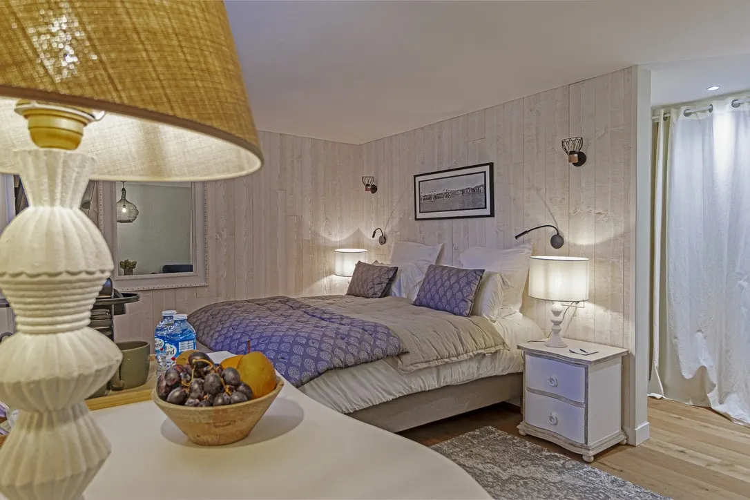 Bedroom at La Plonplonière with a double bed, lit bedside lamps, white panelling and a fruit bowl