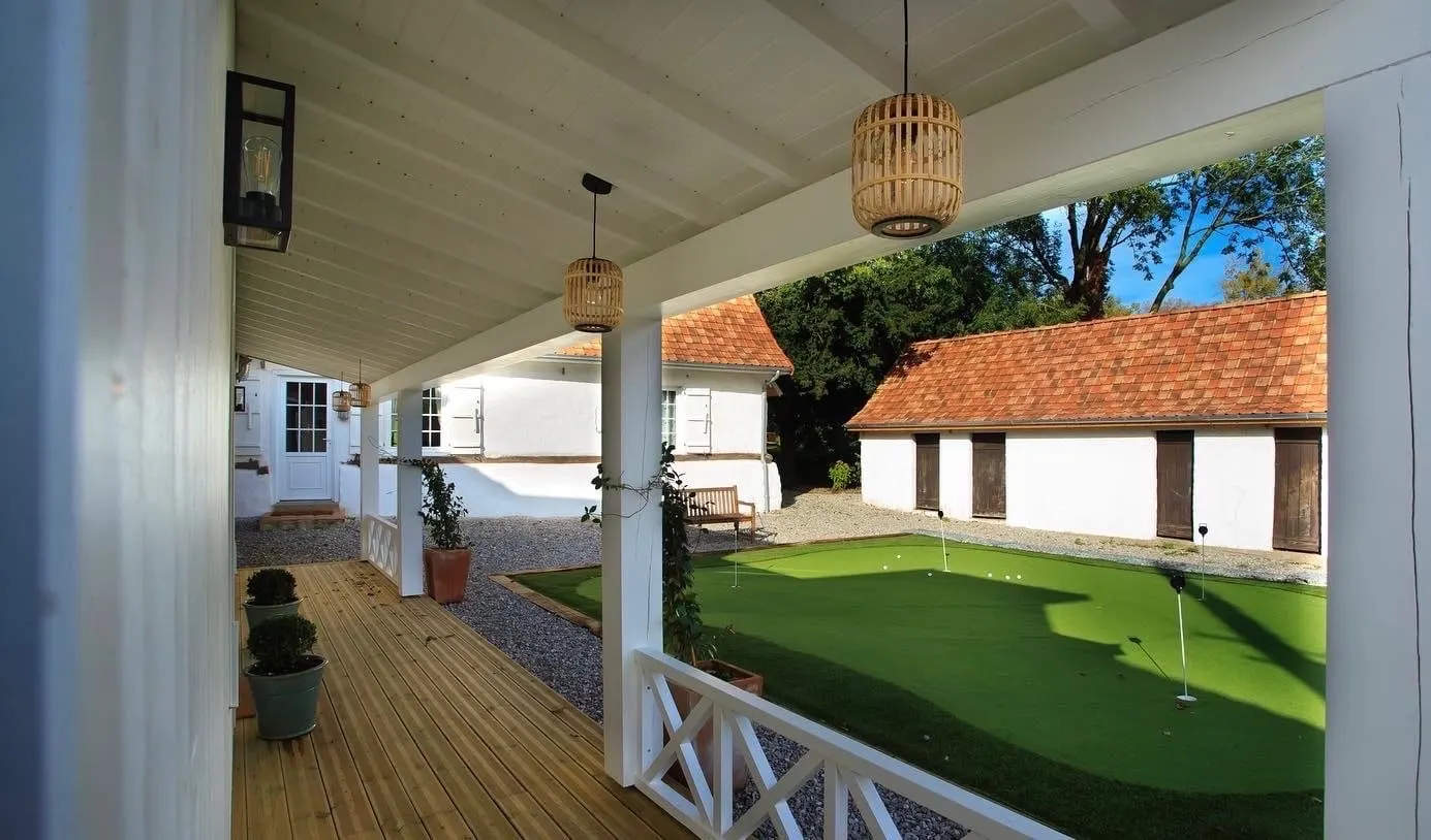 Covered timber veranda at La Plonplonière overlooking a putting green and white outbuildings