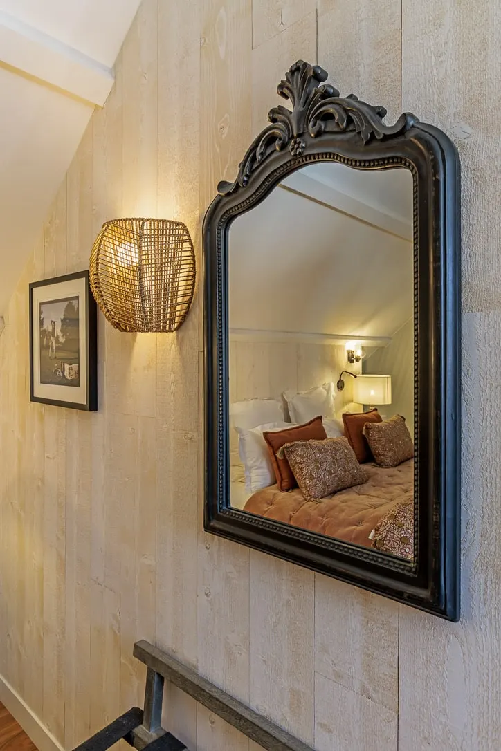 Antique black mirror and rattan wall light on pale panelling, reflecting a bed with russet cushions, La Plonplonière