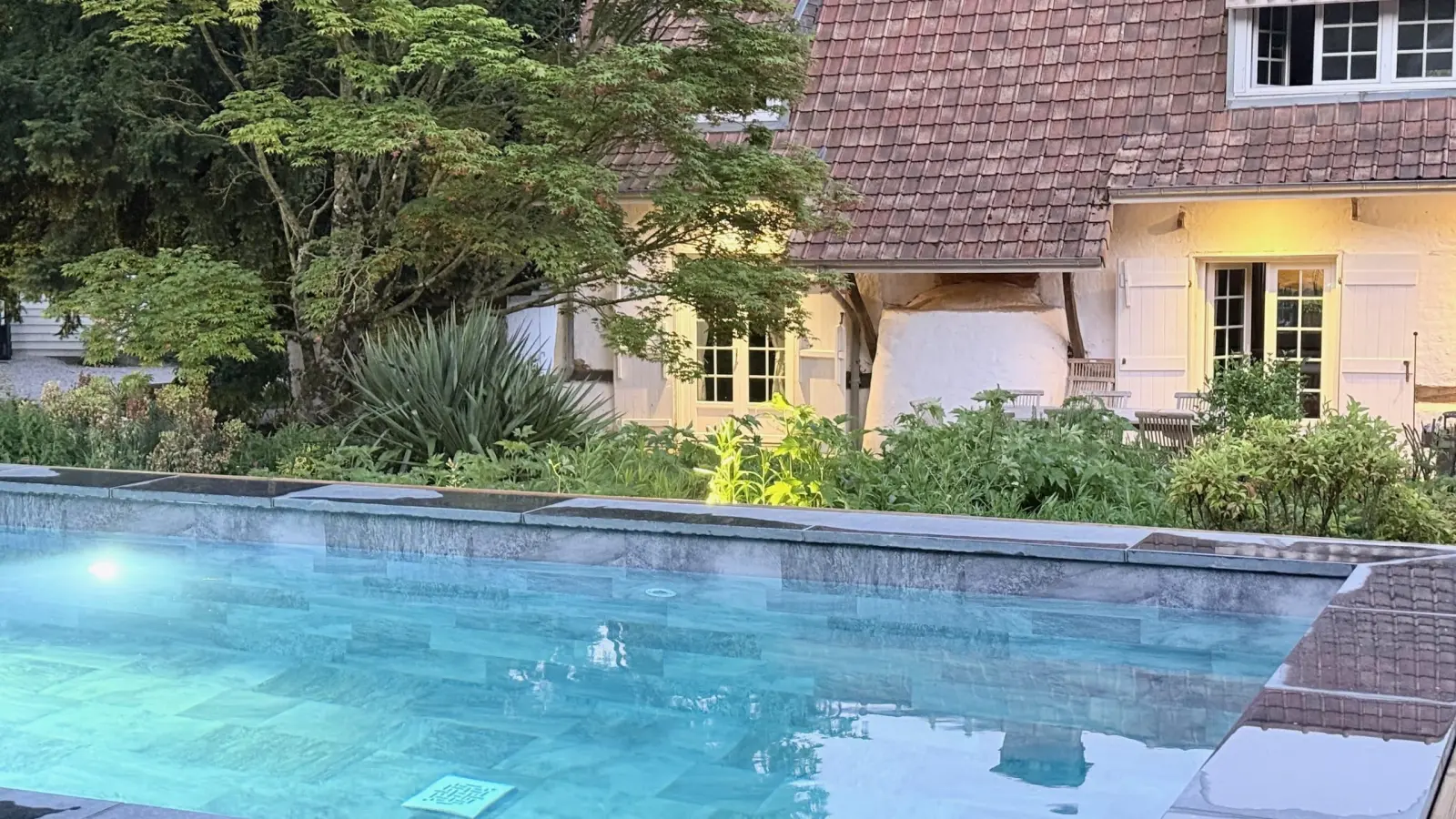 Pool lit at dusk before the house with its brown tiled roof and garden, La Plonplonière