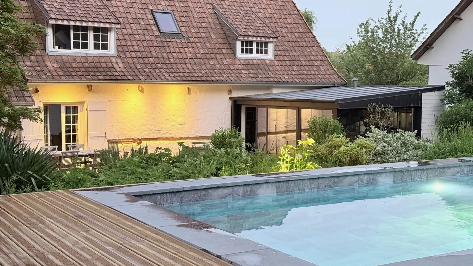 Pool and timber decking before the house lit at dusk, with a tiled roof and dormer windows, La Plonplonière