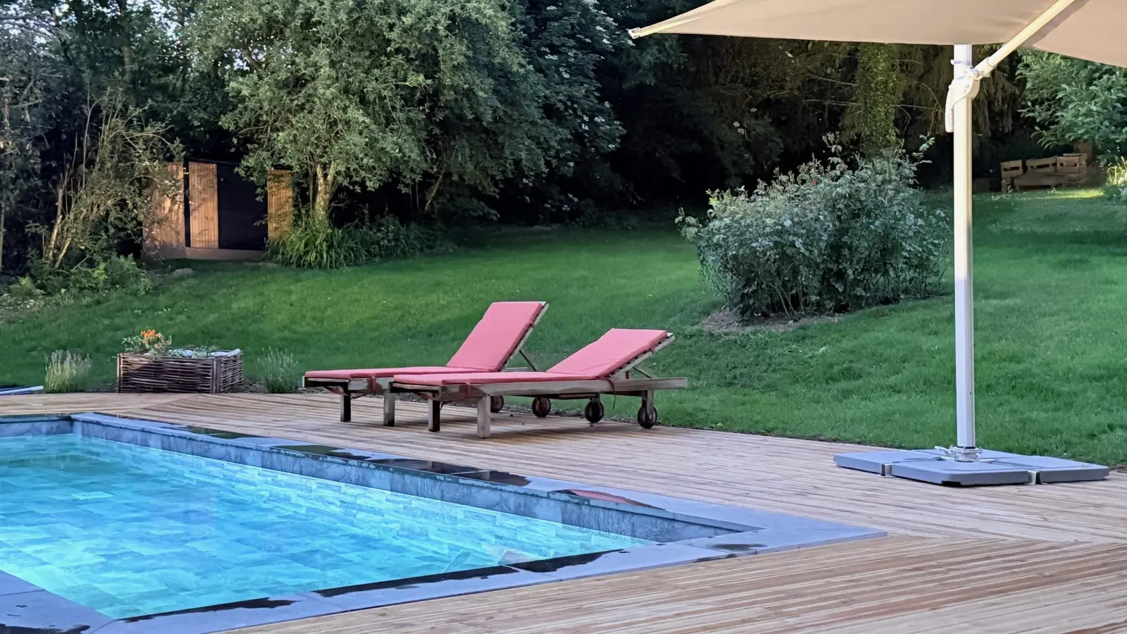 Pool with two red sun loungers and a cantilever parasol on timber decking, lawn and trees, La Plonplonière