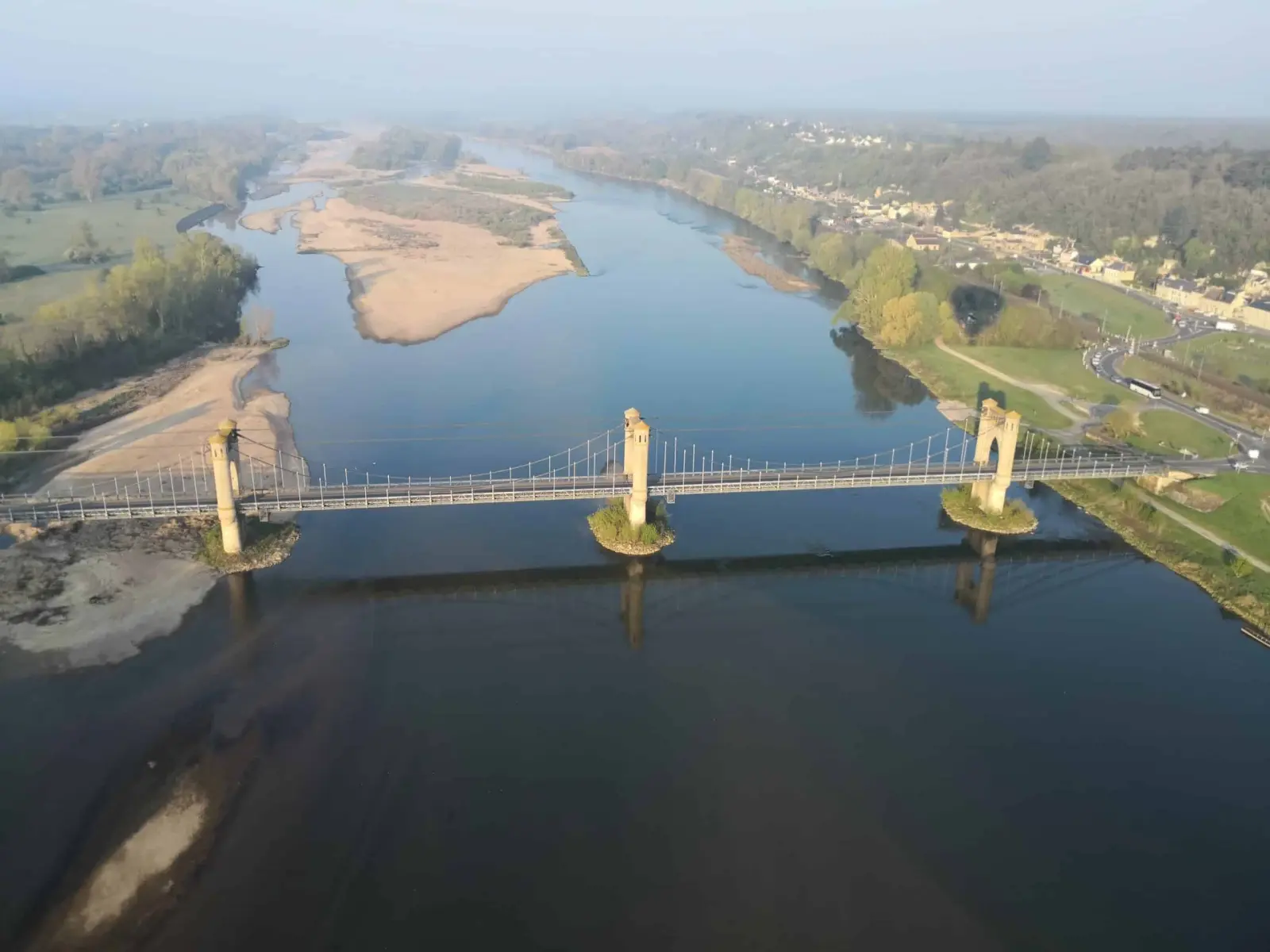 Aerial view of a suspension bridge over a wide river with sandbanks and a small town on the bank