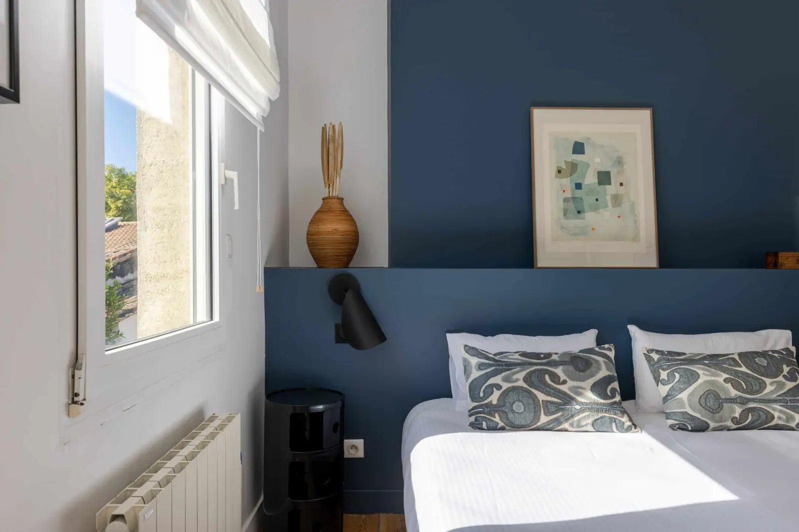 Bedroom with a midnight-blue wall, patterned cushions, black wall lamp and window over the rooftops, Les Séraphines