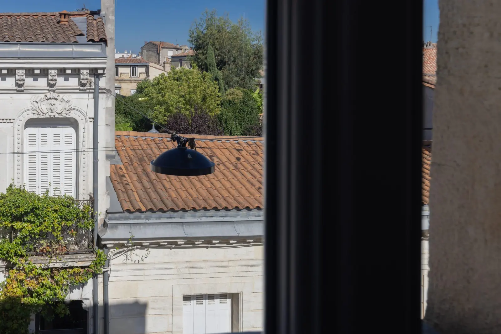 View from a window over tiled rooftops and a stone façade with white shutters, Les Séraphines