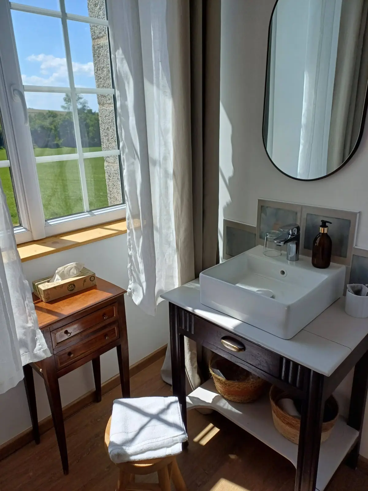Washbasin corner in the Bouli Boula room: white basin, dark wood stand, oval mirror, meadow view, Ty Monde