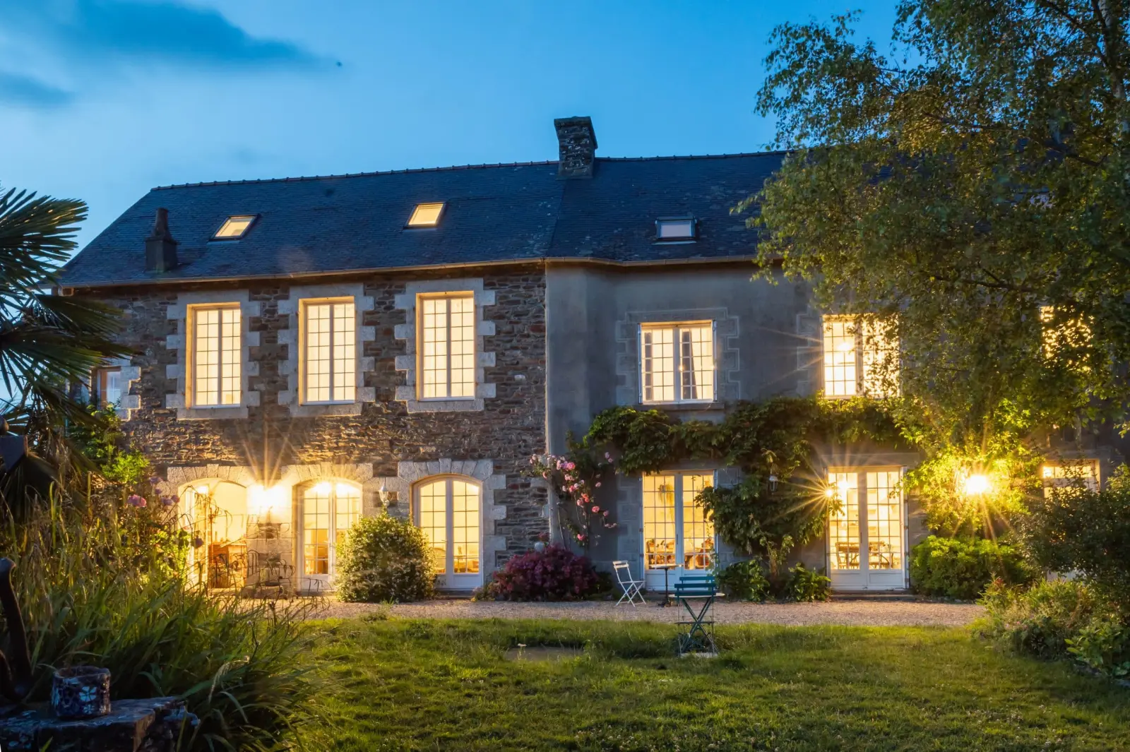 Façade en pierre de Ty Monde éclairée à la tombée du jour, pelouse et jardin au premier plan