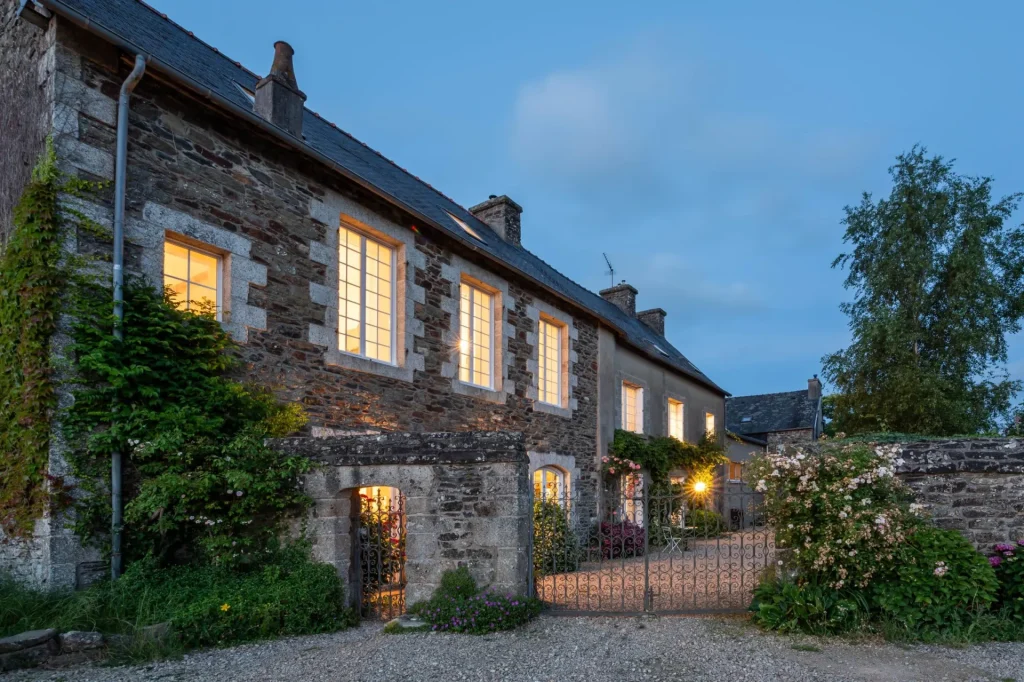 Stone façade of Ty Monde with lit windows at dusk, wrought-iron gate and rose bushes