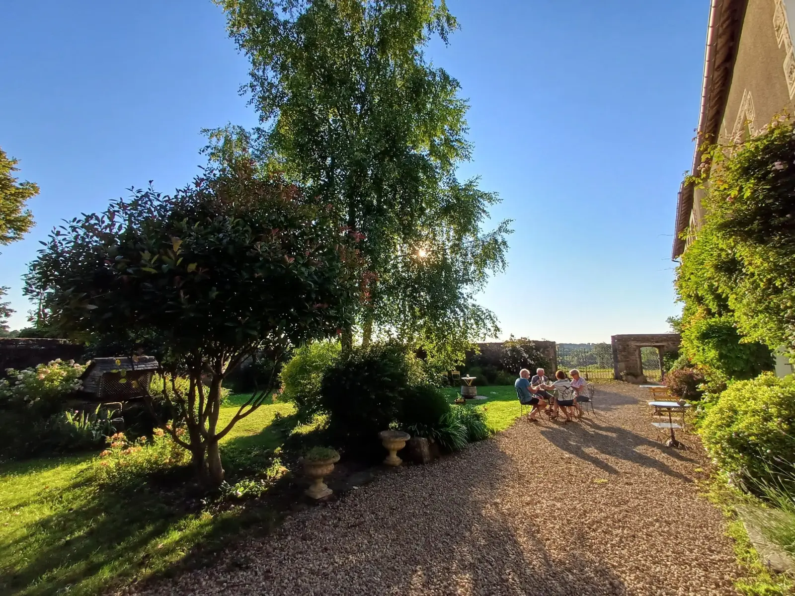 Sunny garden at Ty Monde with a gravel path, tall trees and guests seated at a table